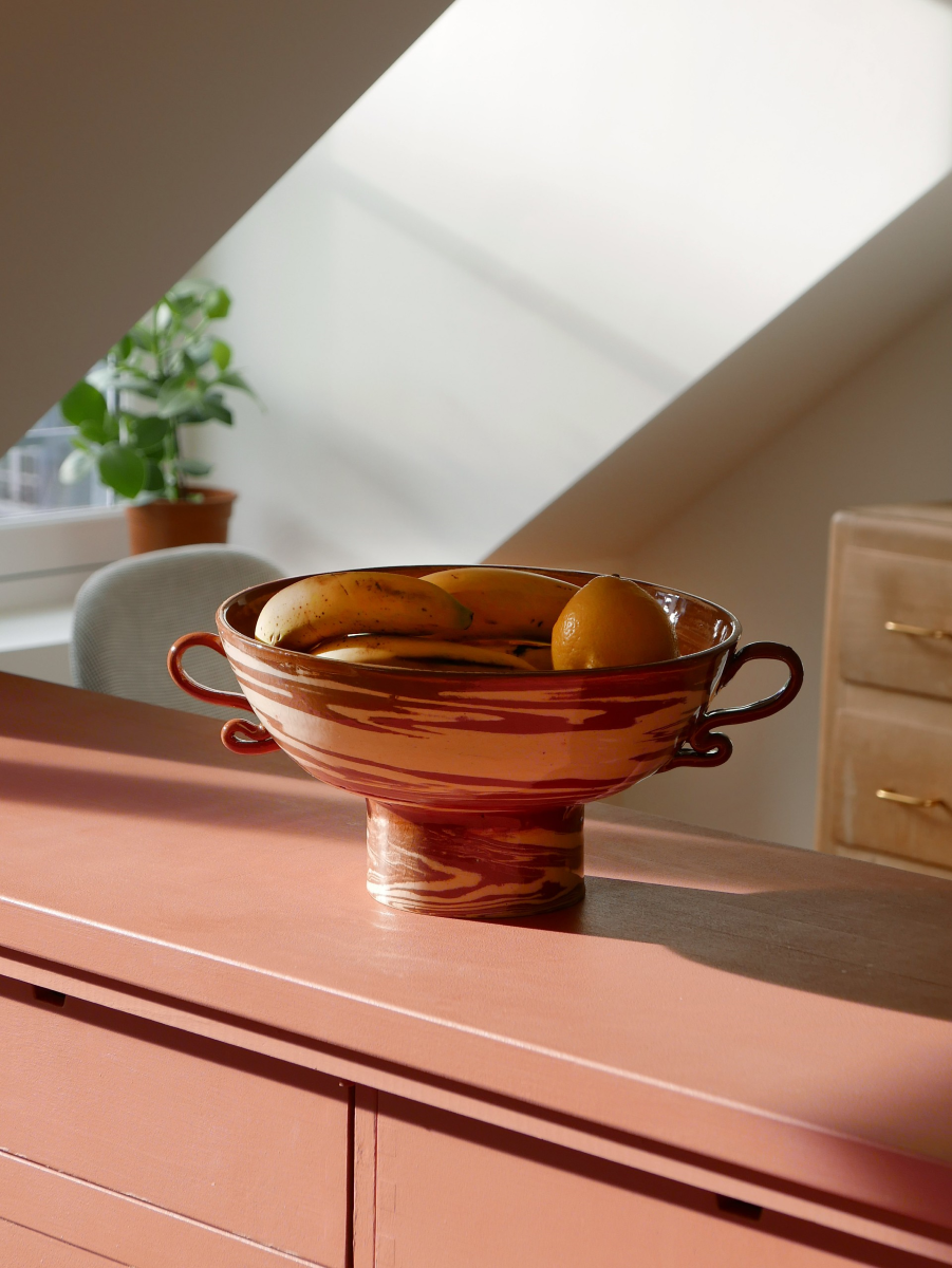 Ces objets en faïence émaillée sont entièrement façonnés à la main par un artisan-potier marocain travaillant dans l’Atlas. Gardien d’une tradition séculaire, il a repris l’atelier familial et perpétue un savoir-faire précieux. Les objets sont tournés un par un à la main en utilisant la technique des terres mêlées. Il s’agit de mélanger des argiles blanches et rouges afin de créer des motifs aléatoires marbrés ou zébrés dans des teintes naturelles terracotta. Chacun de ces objets est ainsi totalement unique