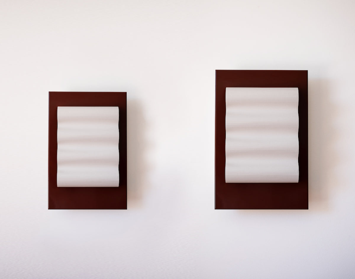 Les appliques Frame Alu Brun de Violaine d'Harcourt sont fabriquées à la main au Portugal, en aluminium laqué brun-rouge et papier.  Dotées d'un design minimaliste et rendant un subtil hommage à l'architecture moderne, les appliques murales Frame sont réalisées avec des matériaux contrastés qui rehaussent le toucher doux d'une vague de papier.