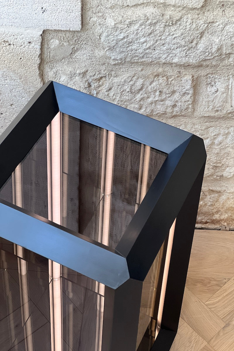 Fabriquée dans une structure en bois teinté noir Napoléon III, pour illustrer son interprétation conceptuelle de l'espace et de la lumière. Le verre sans tain de chaque facette dissimule un ensemble surprenant de miroirs et de lumières. Une fois activé, le rétro-éclairage laisse la magie opérer et le regard captivé pénètre dans un tunnel sans fin.