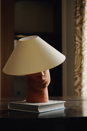 Fabriquée à base de grès orange brut chamotté, la lampe à poser Timide reprend un visage affirmé. Cette pièce fait partie du duo de lampes "Les Sœurs" proposé par l'artiste Thalia Dalecky.