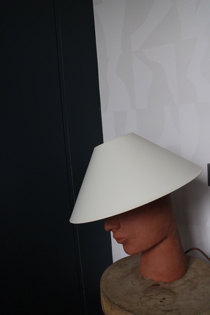 Fabriquée à base de grès orange brut chamotté, la lampe à poser Timide reprend un visage affirmé. Cette pièce fait partie du duo de lampes "Les Sœurs" proposé par l'artiste Thalia Dalecky.