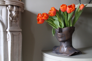 Le vase Tête de Bronze est réalisé par l'artiste Thalia Dalecky avec du grès orange. Sa surface patinée dévoile un visage sculpté dans un grès émaillé noir métallisé.&nbsp; À la lumière, des reflets dorés jouent sur ses contours, ajoutant une touche de mystère à cette pièce d'art captivante, où le bronze robuste rencontre la délicatesse du grès.