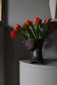 Le vase Tête de Bronze est réalisé par l'artiste Thalia Dalecky avec du grès orange. Sa surface patinée dévoile un visage sculpté dans un grès émaillé noir métallisé.&nbsp; À la lumière, des reflets dorés jouent sur ses contours, ajoutant une touche de mystère à cette pièce d'art captivante, où le bronze robuste rencontre la délicatesse du grès.