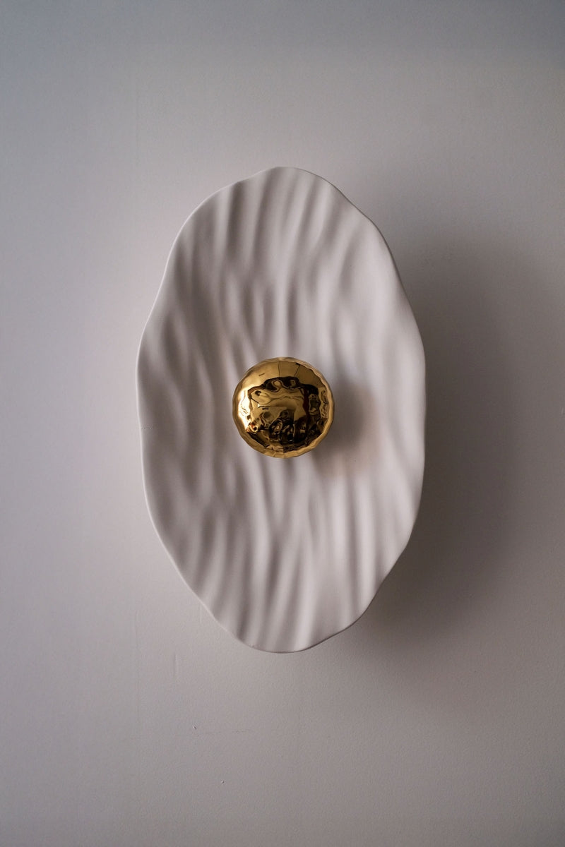 Façonnée à la main, l’applique Vesta associe un disque en faïence émaillée doré 24K, à une structure en plâtre sculpté aux lignes organiques.

Conçue comme une offrande à la lumière, Vesta rend hommage à la déesse du foyer. Elle diffuse une lumière douce et enveloppante, idéale pour instaurer une atmosphère apaisante et sacrée.