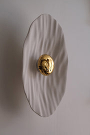 Façonnée à la main, l’applique Vesta associe un disque en faïence émaillée doré 24K, à une structure en plâtre sculpté aux lignes organiques.

Conçue comme une offrande à la lumière, Vesta rend hommage à la déesse du foyer. Elle diffuse une lumière douce et enveloppante, idéale pour instaurer une atmosphère apaisante et sacrée.