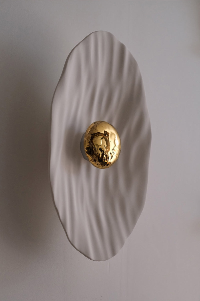 Façonnée à la main, l’applique Vesta associe un disque en faïence émaillée doré 24K, à une structure en plâtre sculpté aux lignes organiques.

Conçue comme une offrande à la lumière, Vesta rend hommage à la déesse du foyer. Elle diffuse une lumière douce et enveloppante, idéale pour instaurer une atmosphère apaisante et sacrée.